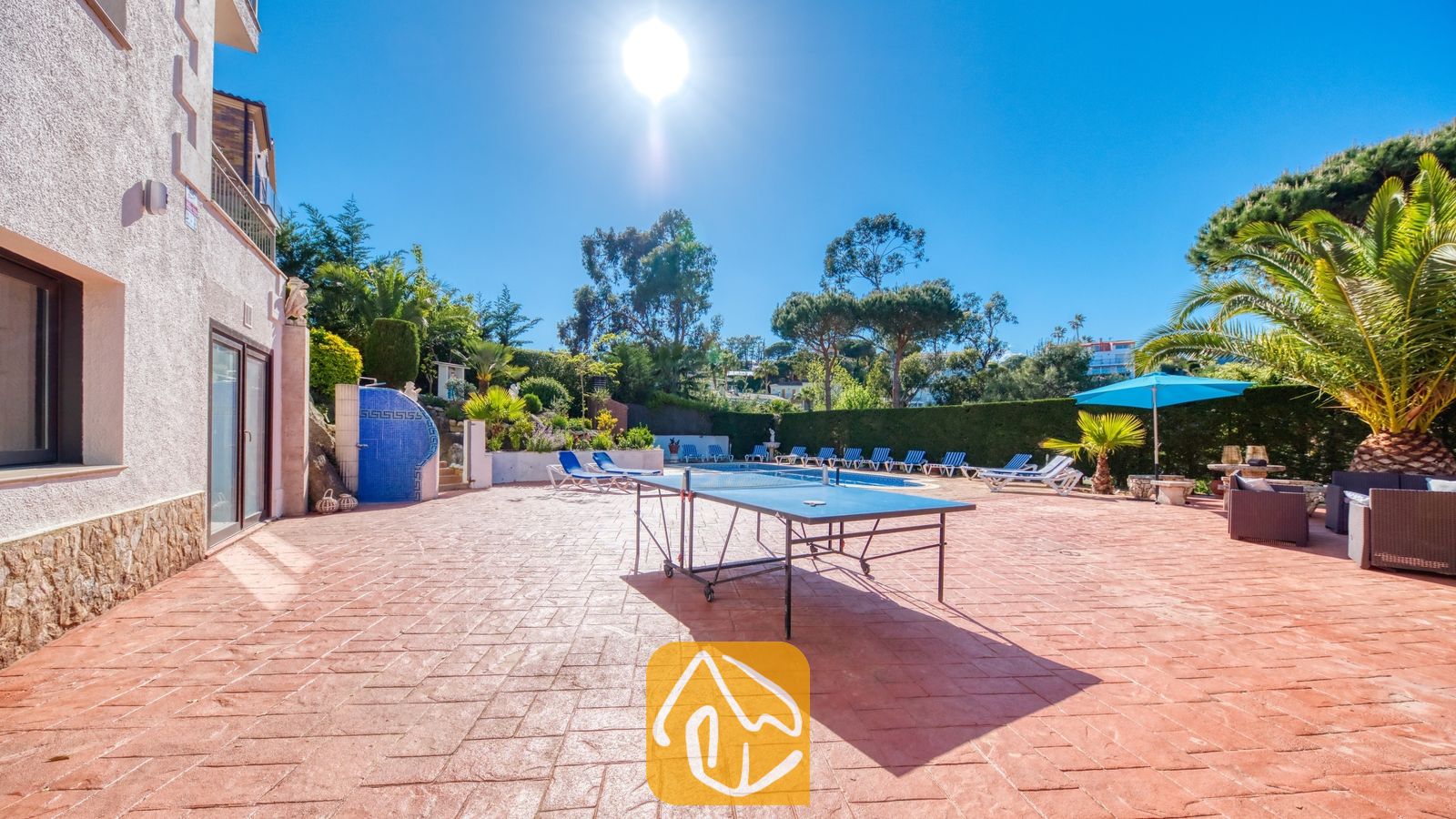 Vakantievilla's Costa Brava Spanje - Villa Jaruco - Zonnig terras bij Villa Jaruco met een blauwe pingpongvloer op een terracotta printvloer, met een zwembad en ligstoelen op de achtergrond.