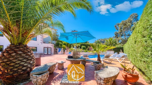 Vakantievilla's Costa Brava Spanje - Villa Jaruco - Zonnig terras bij Villa Jaruco, voorzien van een stenen tafel met uitgesneden banken onder een helderblauwe parasol, omringd door palmbomen en een zwembad.