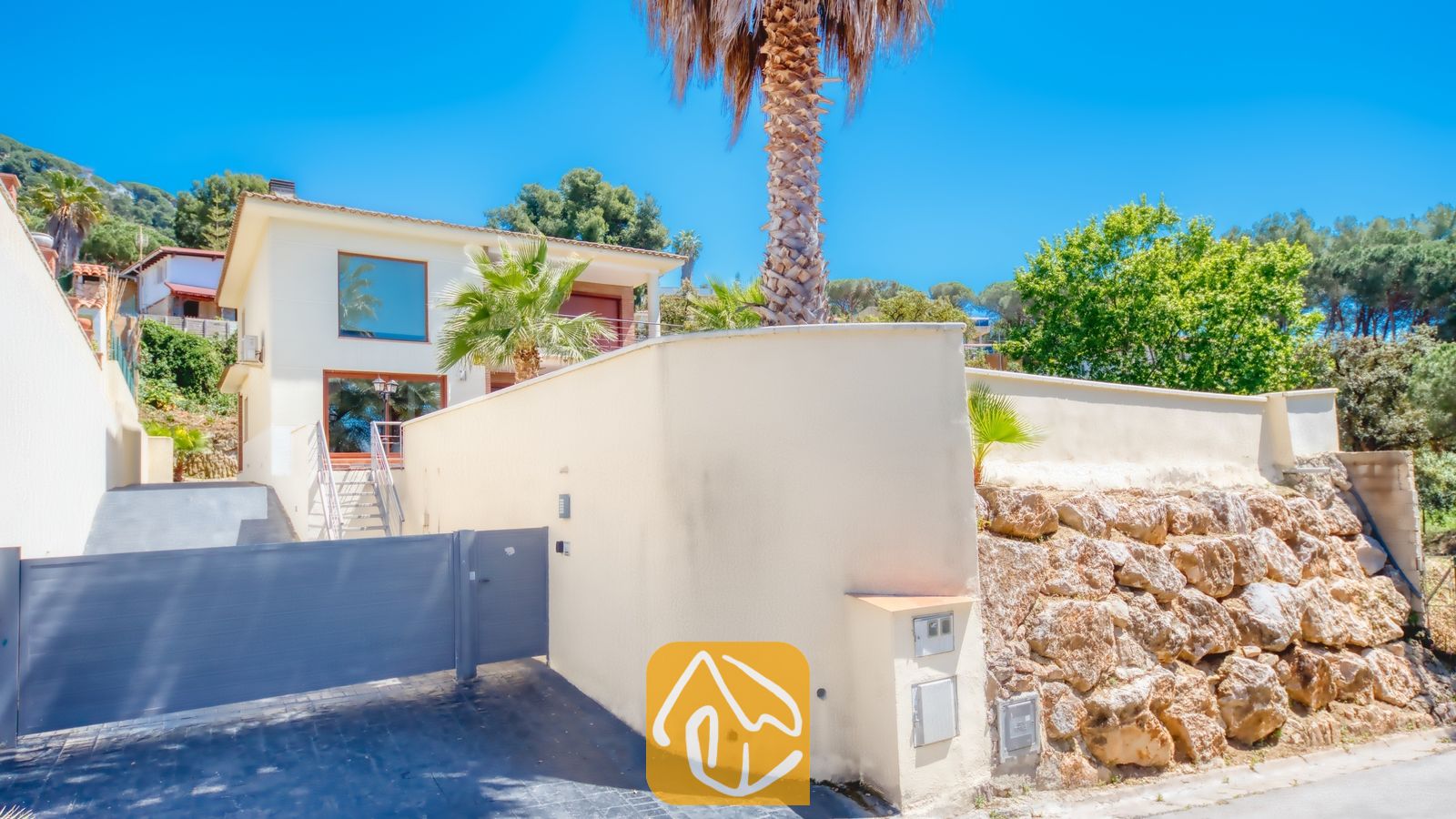 Vakantievilla's Costa Brava Spanje - Villa Marcella - Buitenaanzicht van Villa Marcella met een wit huis van twee verdiepingen achter een hoge erfafscheiding, een grijze toegangspoort, palmbomen en een stenen keermuur onder een blauwe lucht.