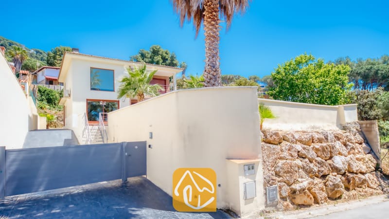 Villas de vacances Costa Brava Espagne - Villa Marcella - Vue extérieure de la Villa Marcella présentant une maison blanche à deux étages derrière un mur d'enceinte élevé, un portail d'entrée gris, des palmiers et un mur de soutènement en pierre sous un ciel bleu.