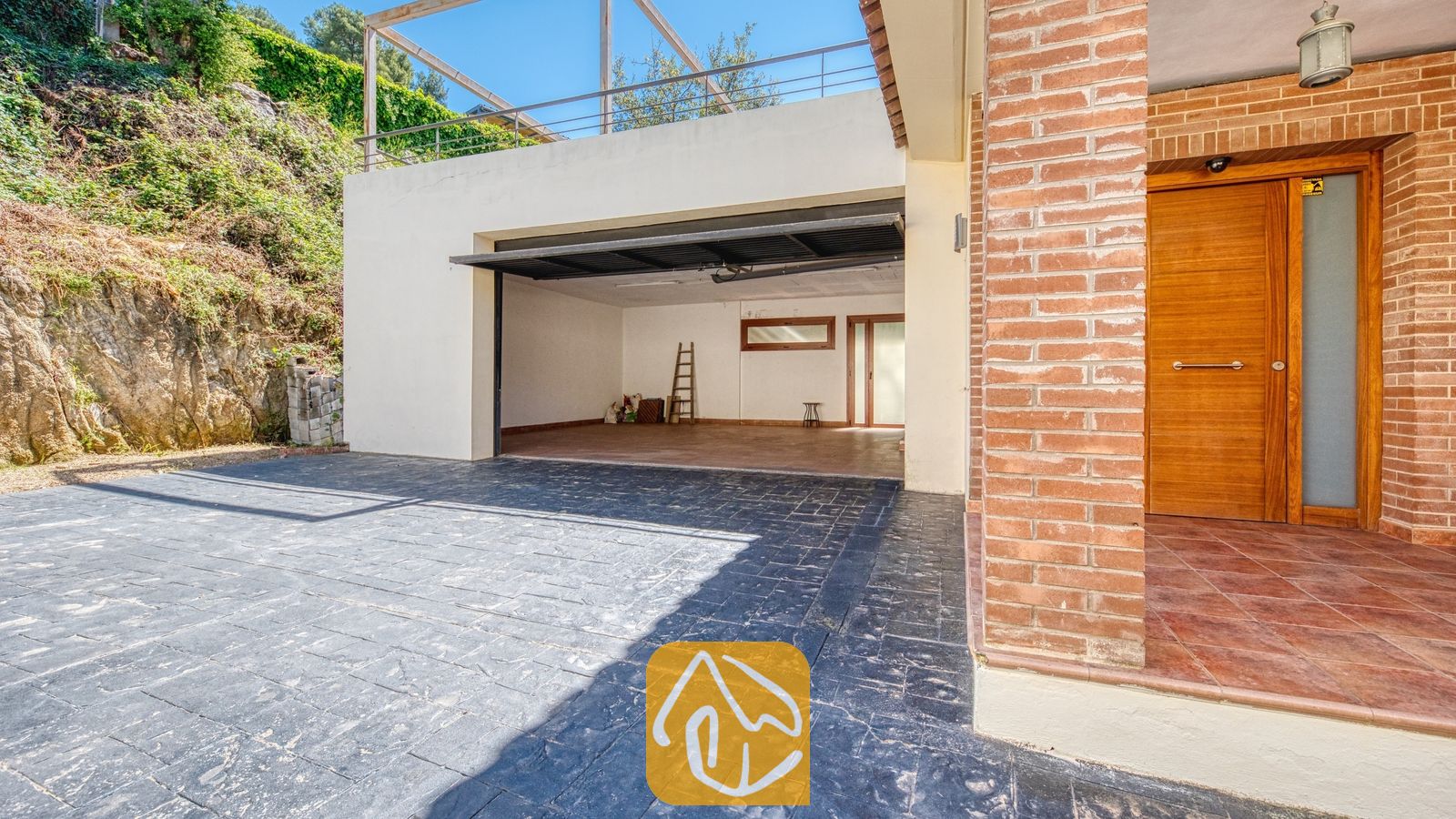 Vakantievilla's Costa Brava Spanje - Villa Marcella - De oprit en open garage van Villa Marcella, voorzien van m Outsourced bestrating van donker asfalt/beton en een entree met rode bakstenen en een houten voordeur.
