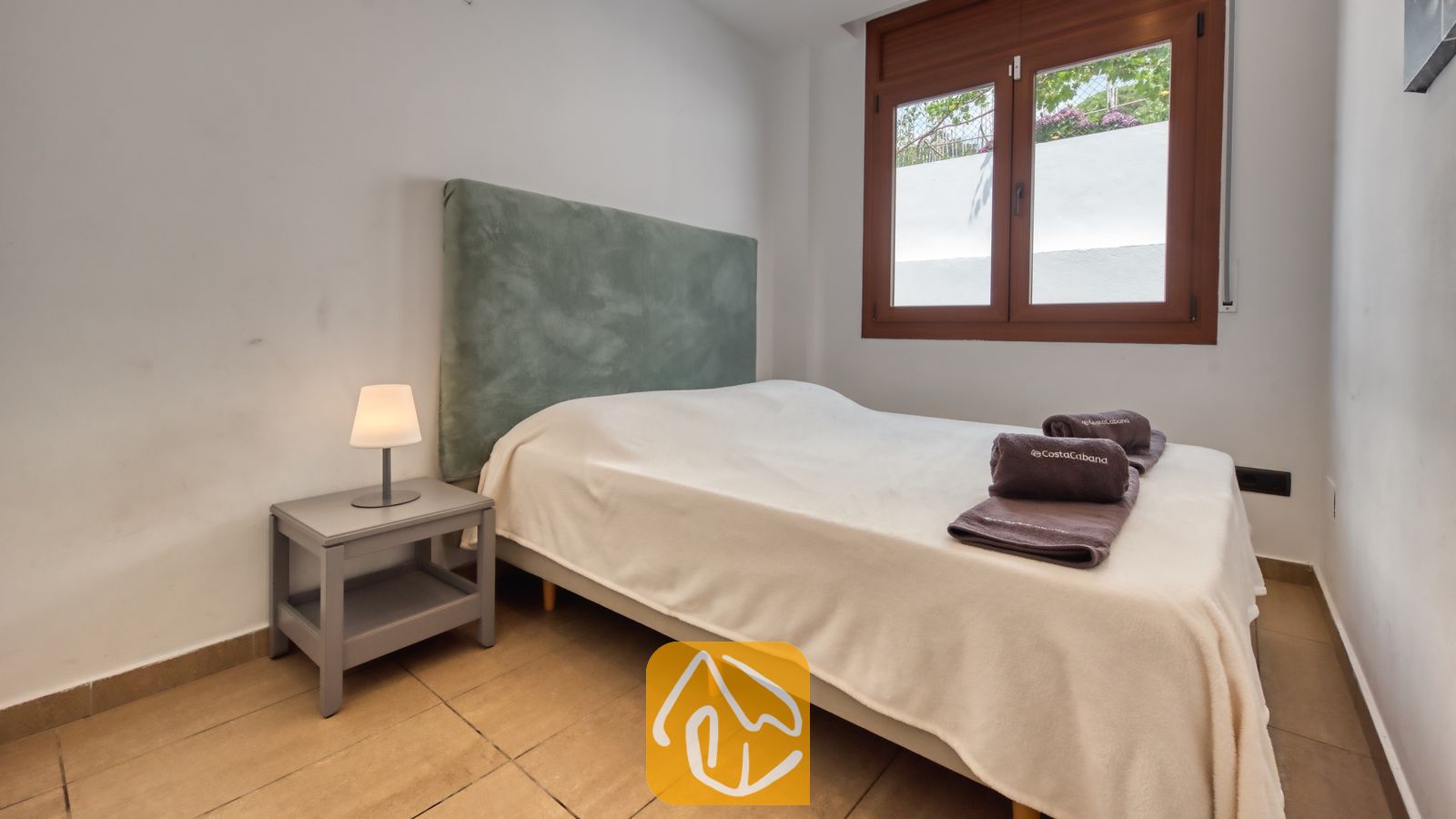 Vakantievilla's Costa Brava Spanje - Villa Marcella - Slaapkamer met een tweepersoonsbed voorzien van een gedempt groen hoofdbord, witter beddengoed, opgevouwen handdoeken, een grijs nachtkastje met een klein lampje en een bruin houten raamkozijn.