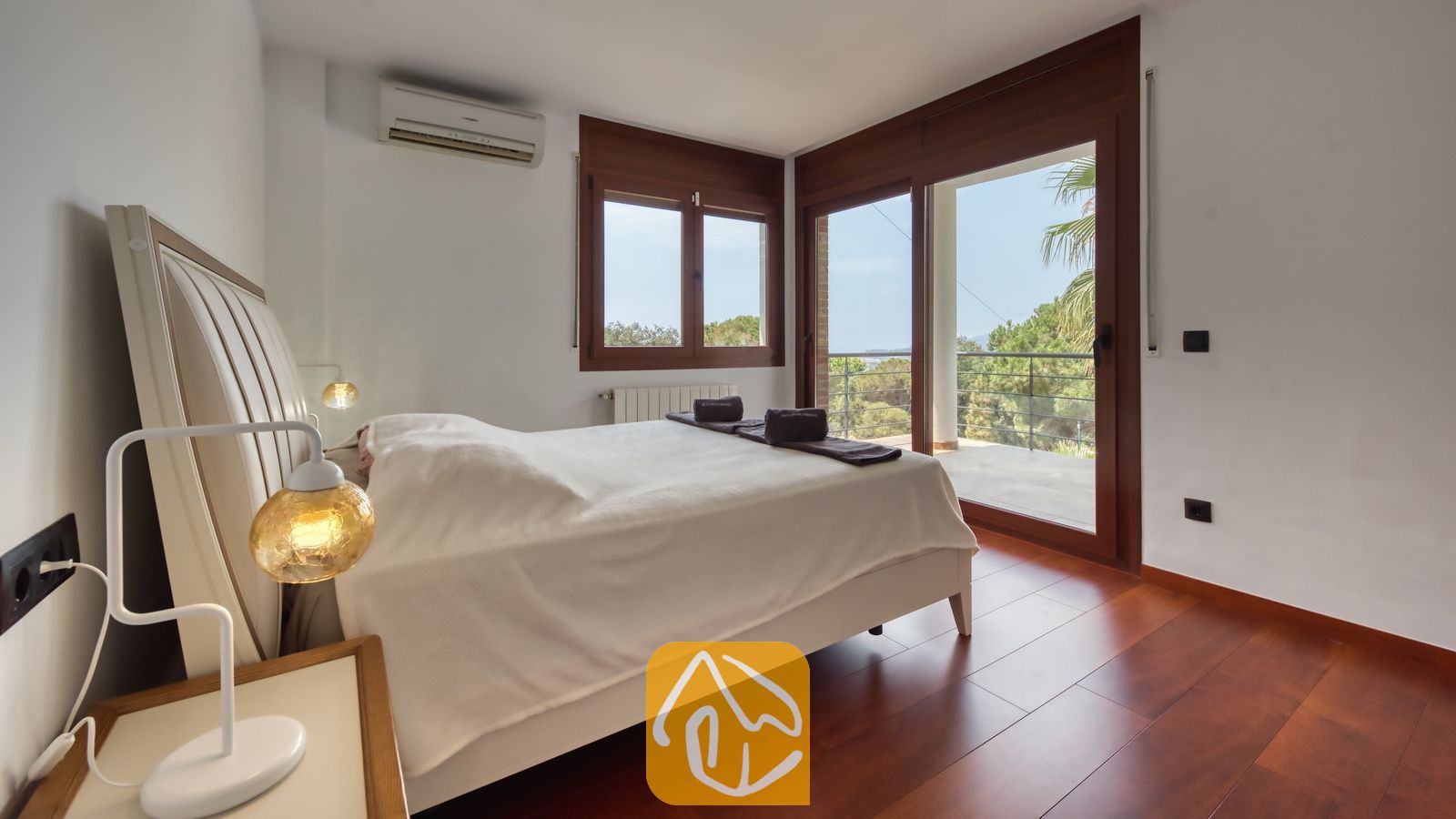 Vakantievilla's Costa Brava Spanje - Villa Marcella - Slaapkamer met een wit tweepersoonsbed, donkere houten vloer en glazen schuifdeuren die uitkomen op een buitenbalkon bij Villa Marcella.