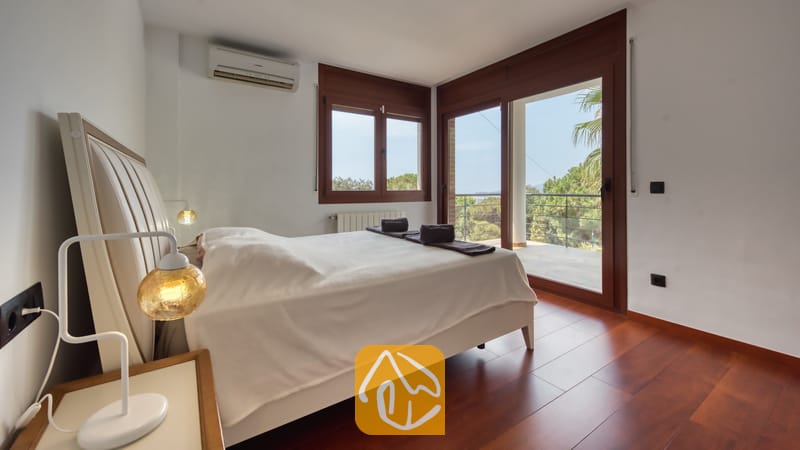 Villas de vacances Costa Brava Espagne - Villa Marcella - Chambre à coucher dotée d'un lit double blanc, de parquets en bois sombre et de portes coulissantes en verre s'ouvrant sur un balcon extérieur à la Villa Marcella.