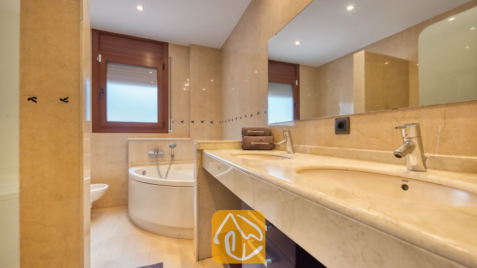 Vakantievilla's Costa Brava Spanje - Villa Marcella - Badkamer met een marmeren dubbele wastafel, een hoekbad onder een houten raam en beige tegelmuren.