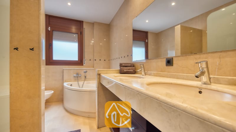 Villas de vacances Costa Brava Espagne - Villa Marcella - Salle de bain comprenant une double vasque en marbre, une baignoire d'angle sous une fenêtre en bois et des murs carrelés de beige.