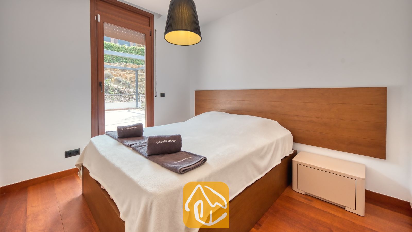 Vakantievilla's Costa Brava Spanje - Villa Marcella - Slaapkamer met een houten hoofdbord, een tweepersoonsbed met opgevouwen grijze handdoeken, een donkere hanglamp en een glazen deur die uitkomt op een buitenterras.