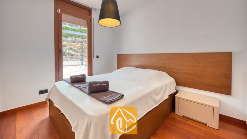 Villas de vacances Costa Brava Espagne - Villa Marcella - Chambre avec une tête de lit en bois, lit double agrémenté de serviettes grises pliées, suspension sombre et porte vitrée donnant sur un patio extérieur.