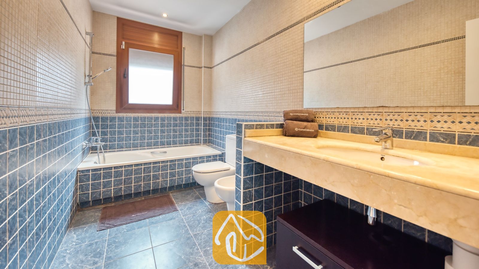 Vakantievilla's Costa Brava Spanje - Villa Marcella - Badkamer met blauw- en beige-mozaïektegels op de wanden, een ligbad onder een houten raamkozijn, een toilet, een bidet en een wastafel met marmeren blad en een grote spiegel.