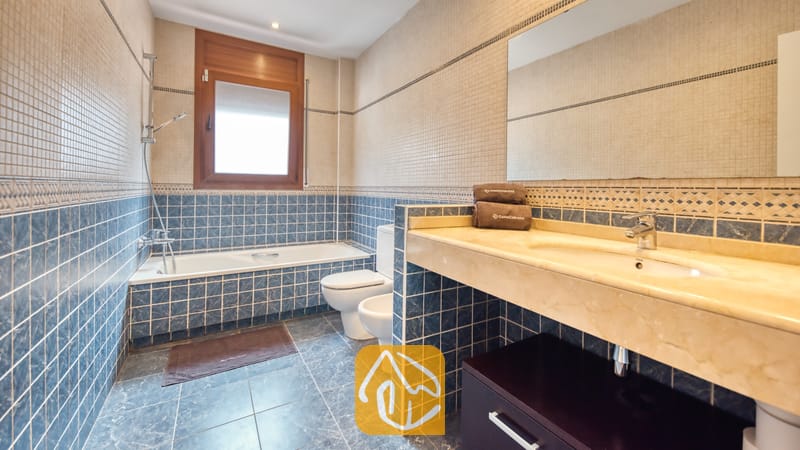 Villas de vacances Costa Brava Espagne - Villa Marcella - Salle de bain dotée de murs ornés de mosaïques bleues et beiges, d'une baignoire située sous une fenêtre à cadre en bois, de toilettes, d'un bidet et d'un meuble sous-vasque en marbre surmonté d'un grand miroir.