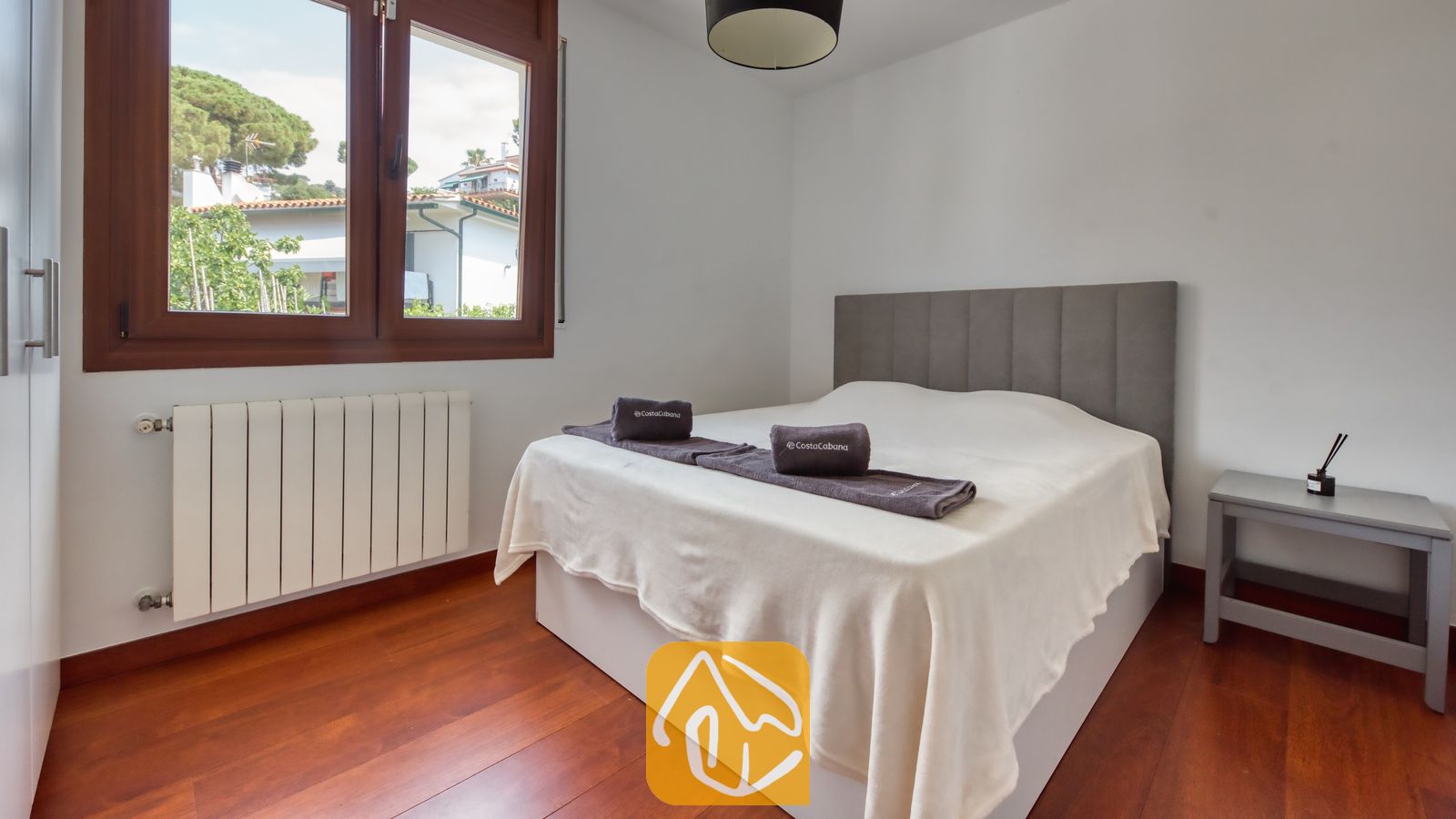 Vakantievilla's Costa Brava Spanje - Villa Marcella - Slaapkamer met een tweepersoonsbed, donkergrijs gestoffeerd hoofdeinde, opgevouwen handdoeken, houten vloer en een raam met uitzicht naar buiten.