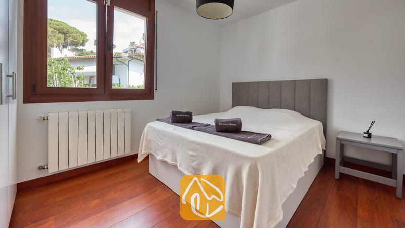 Villas de vacances Costa Brava Espagne - Villa Marcella - Chambre avec un lit double, une tête de lit capitonnée gris foncé, des serviettes pliées, un parquet en bois et une fenêtre offrant une vue sur l'extérieur.