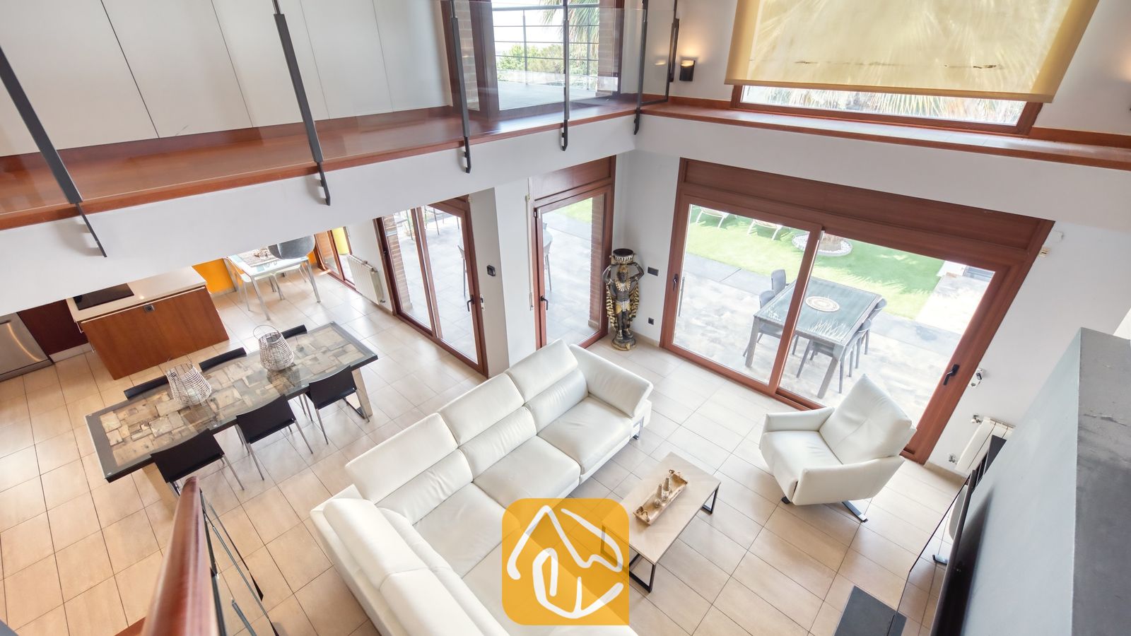Vakantievilla's Costa Brava Spanje - Villa Marcella - Hoogperspectief van de woonkamer in Villa Marcella met een witte, L-vormige leren bank, een bijpassende fauteuil en schuifpui die toegang biedt tot het terras.