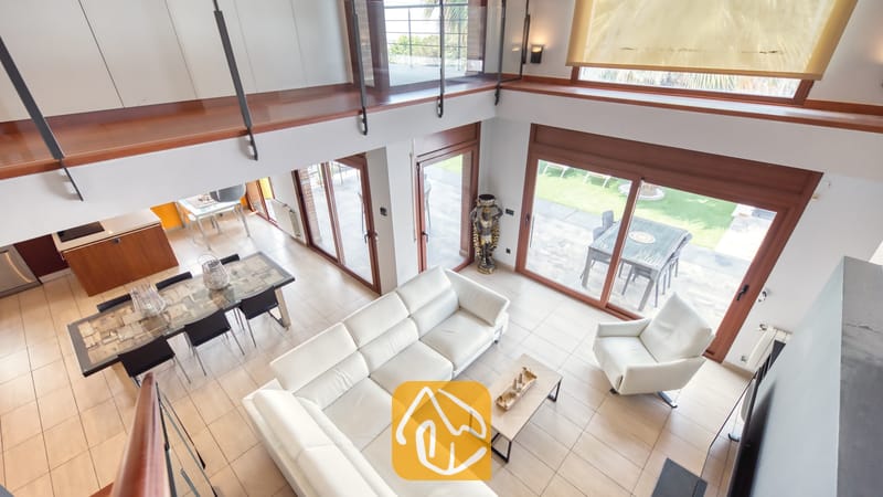 Vakantievilla's Costa Brava Spanje - Villa Marcella - Hoogperspectief van de woonkamer in Villa Marcella met een witte, L-vormige leren bank, een bijpassende fauteuil en schuifpui die toegang biedt tot het terras.