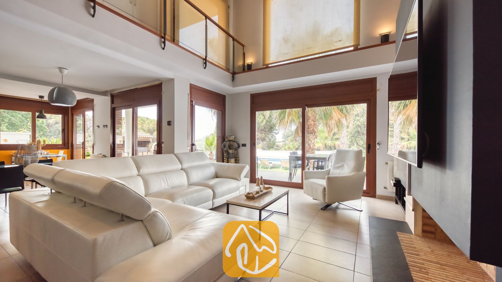 Vakantievilla's Costa Brava Spanje - Villa Marcella - Ruime woonkamer in Villa Marcella met een groot wit leren hoekbankstel, een bijpassende fauteuil, een open haard en glazen schuifpui die toegang biedt tot een terras met palmbomen.