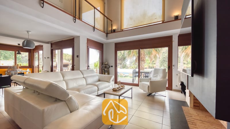 Vakantievilla's Costa Brava Spanje - Villa Marcella - Ruime woonkamer in Villa Marcella met een groot wit leren hoekbankstel, een bijpassende fauteuil, een open haard en glazen schuifpui die toegang biedt tot een terras met palmbomen.