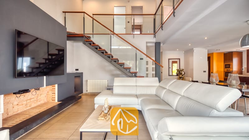 Vakantievilla's Costa Brava Spanje - Villa Marcella - Ruime, open woonkamer in Villa Marcella met een witte lederen hoekbank, een moderne hangende haard, een aan de muur bevestigde televisie en een trap met glazen leuning die naar een overloop op de verdieping leidt.