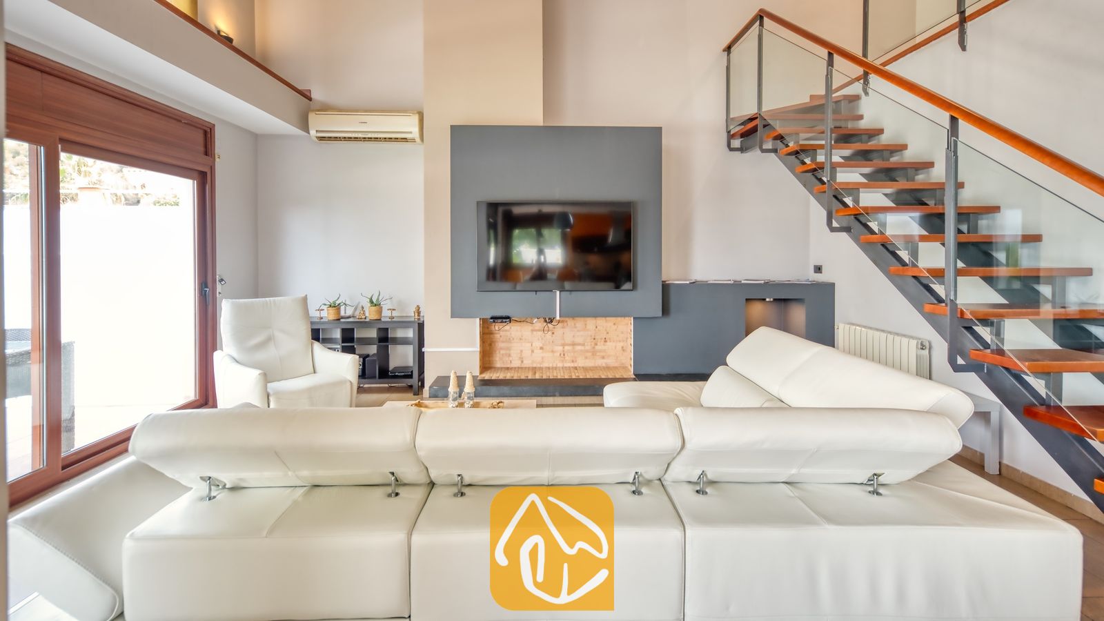 Vakantievilla's Costa Brava Spanje - Villa Marcella - Ruime woonkamer in Villa Marcella, voorzien van een witte L-vormige lederen bank, een flatscreen-televisie aan de muur en een aangesloten houten zwevende trap met glazen leuningen.