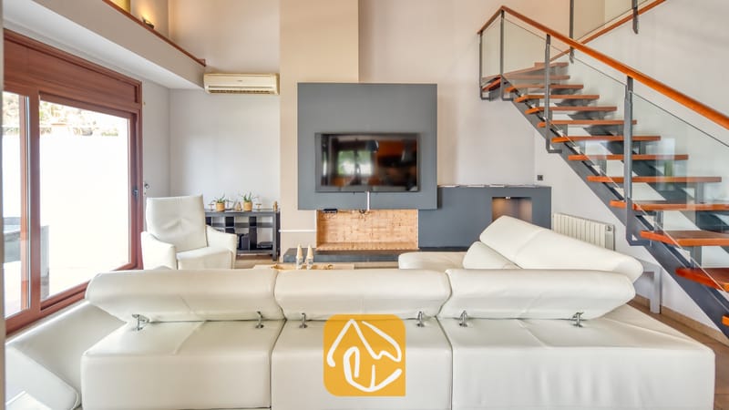 Vakantievilla's Costa Brava Spanje - Villa Marcella - Ruime woonkamer in Villa Marcella, voorzien van een witte L-vormige lederen bank, een flatscreen-televisie aan de muur en een aangesloten houten zwevende trap met glazen leuningen.