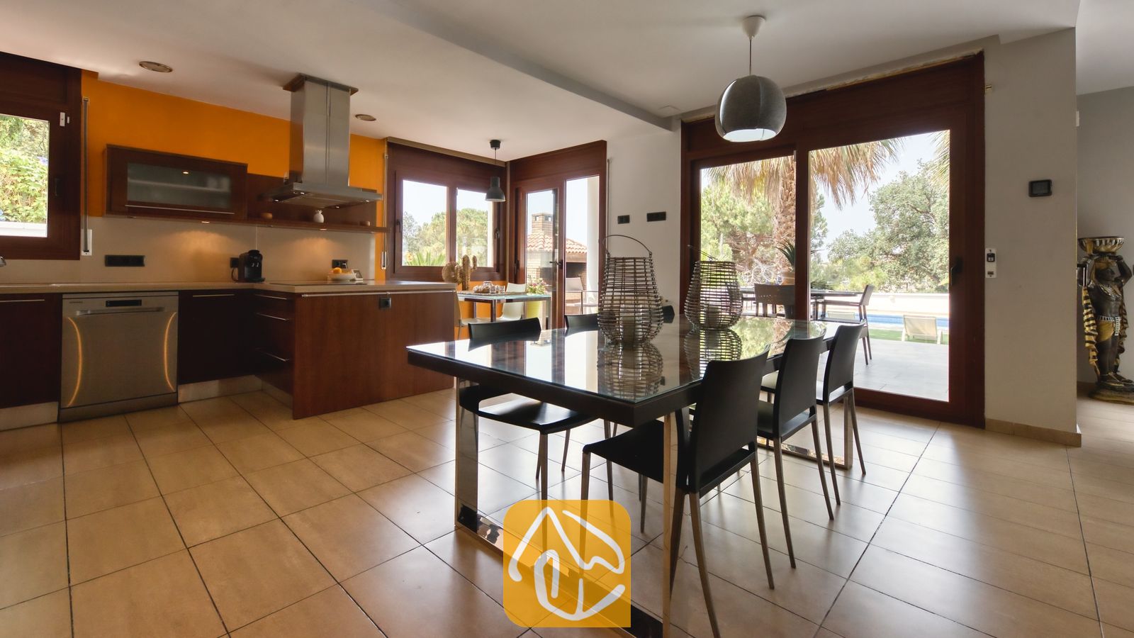 Vakantievilla's Costa Brava Spanje - Villa Marcella - De open eethoek en keuken van Villa Marcella zijn voorzien van een glazen eettafel met zwarte stoelen, een tegelvloer en schuifpui die toegang biedt tot een buitenterras.