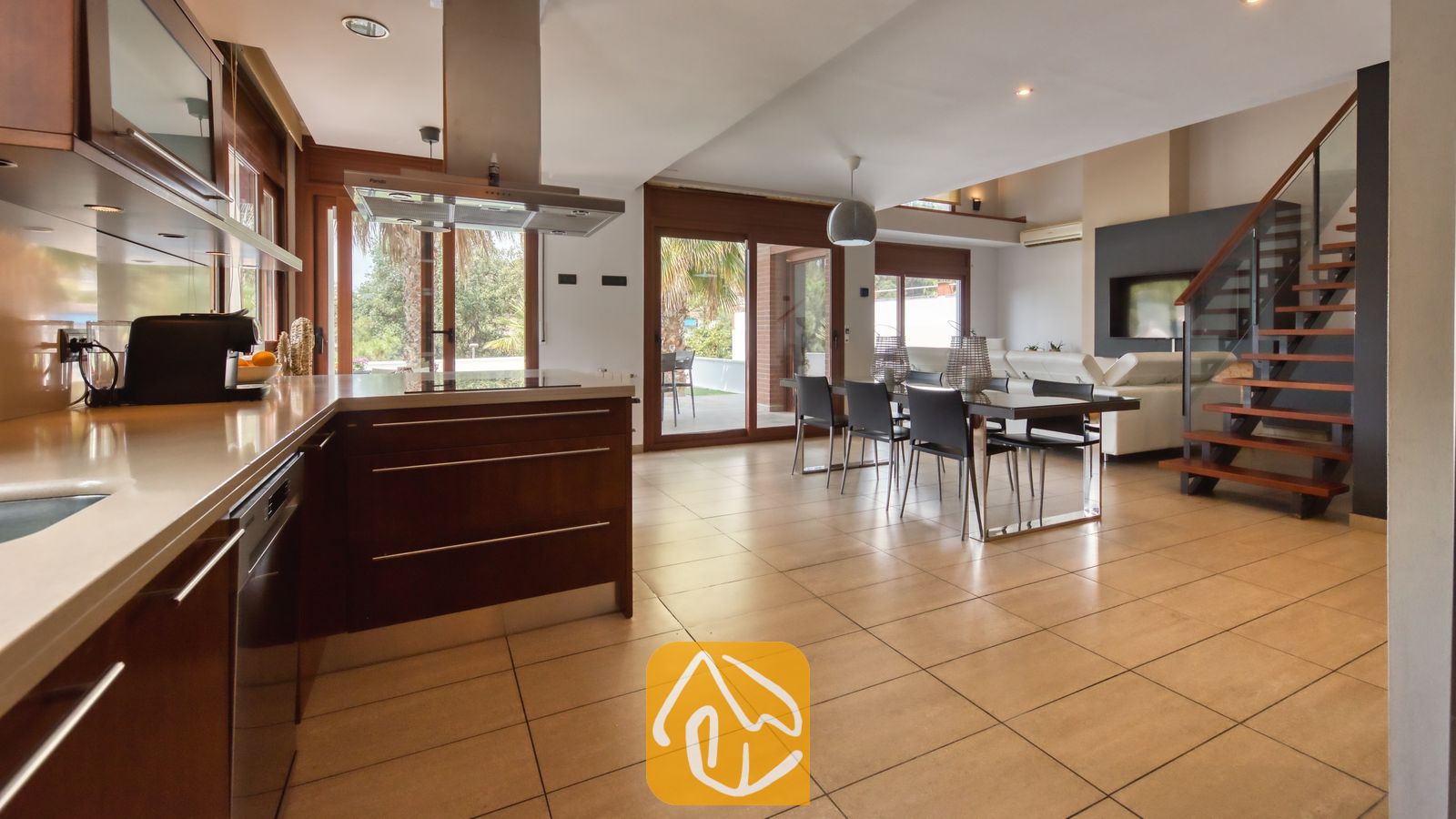 Vakantievilla's Costa Brava Spanje - Villa Marcella - Open-plan keuken en eethoek in Villa Marcella, voorzien van donker houten kasten, een tegelvloer en een houten trap met een glazen balustrade.
