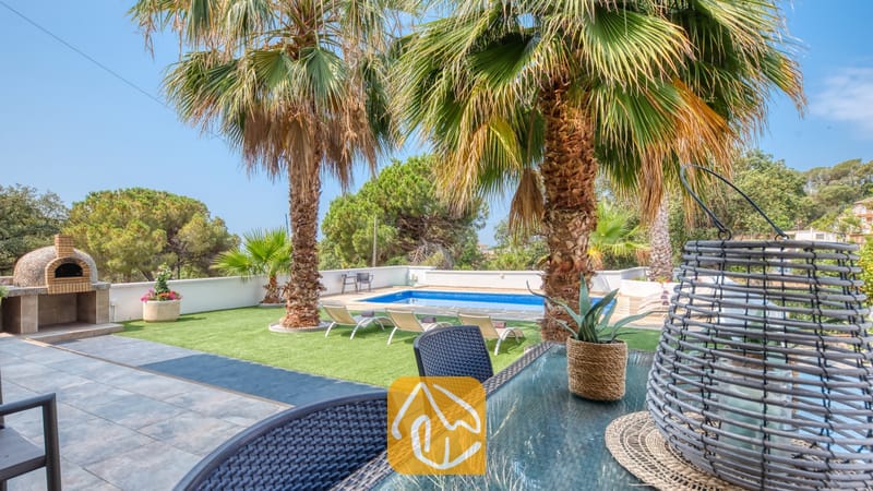 Vakantievilla's Costa Brava Spanje - Villa Marcella - Buitenkant van Villa Marcella met op de voorgrond een glazen eettafel met grijze Wicker stoelen, uitkijkend op een zwembad, palmbomen en een stenen buitenoven.