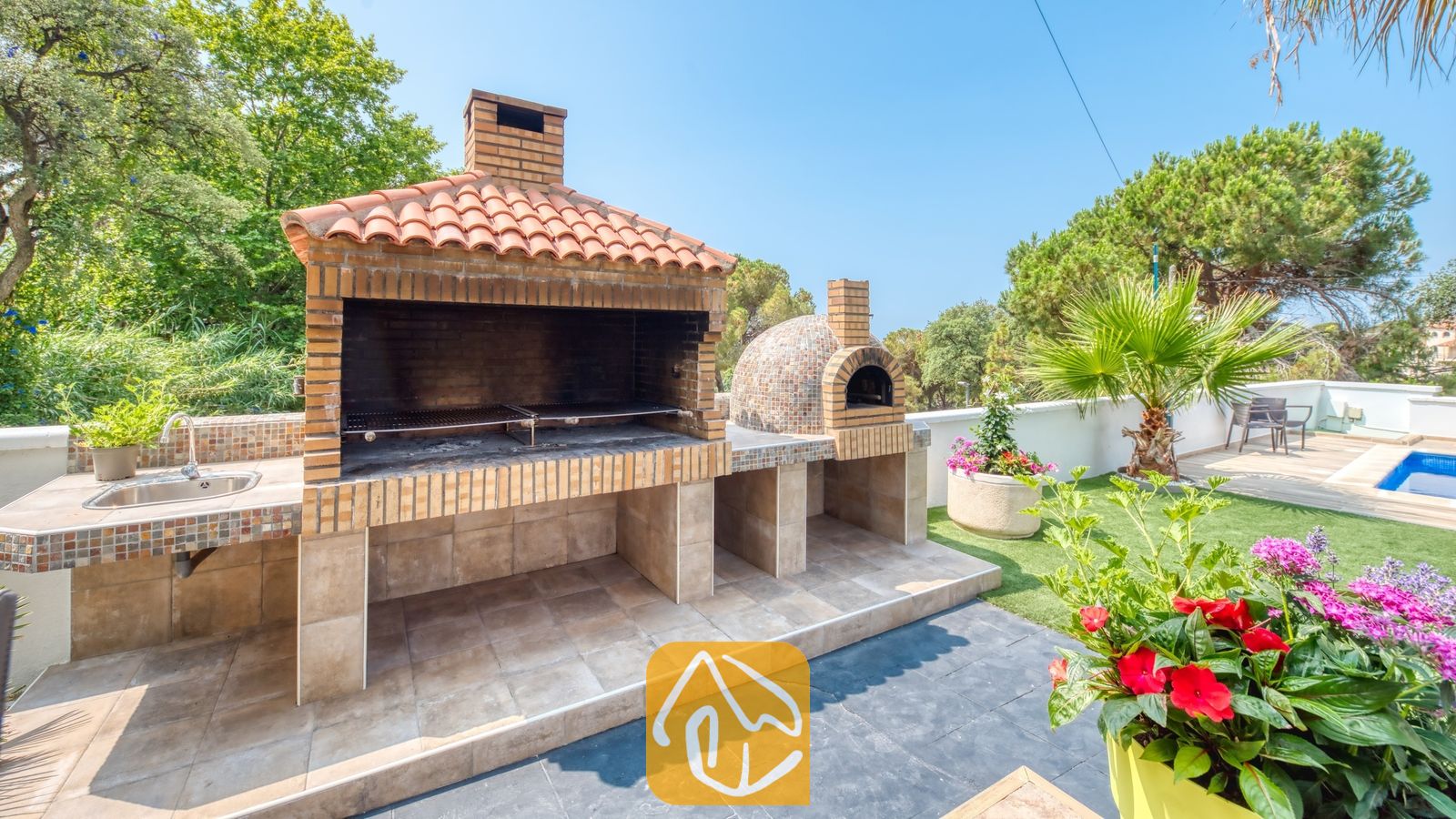 Vakantievilla's Costa Brava Spanje - Villa Marcella - Buitenkeuken met stenen barbecue bij Villa Marcella, voorzien van een pannendak, aangrenzende pizzaoven, ingebouwde gootsteen en een omringend tuinterras.