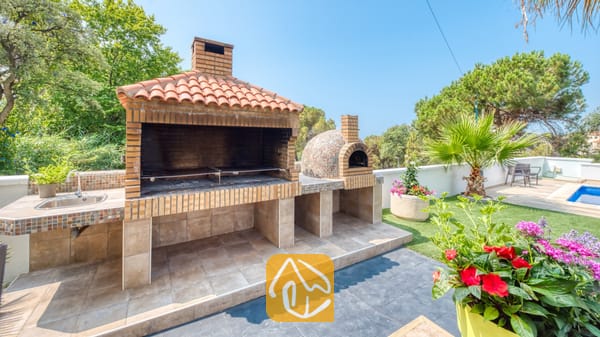 Vakantievilla's Costa Brava Spanje - Villa Marcella - Buitenkeuken met stenen barbecue bij Villa Marcella, voorzien van een pannendak, aangrenzende pizzaoven, ingebouwde gootsteen en een omringend tuinterras.