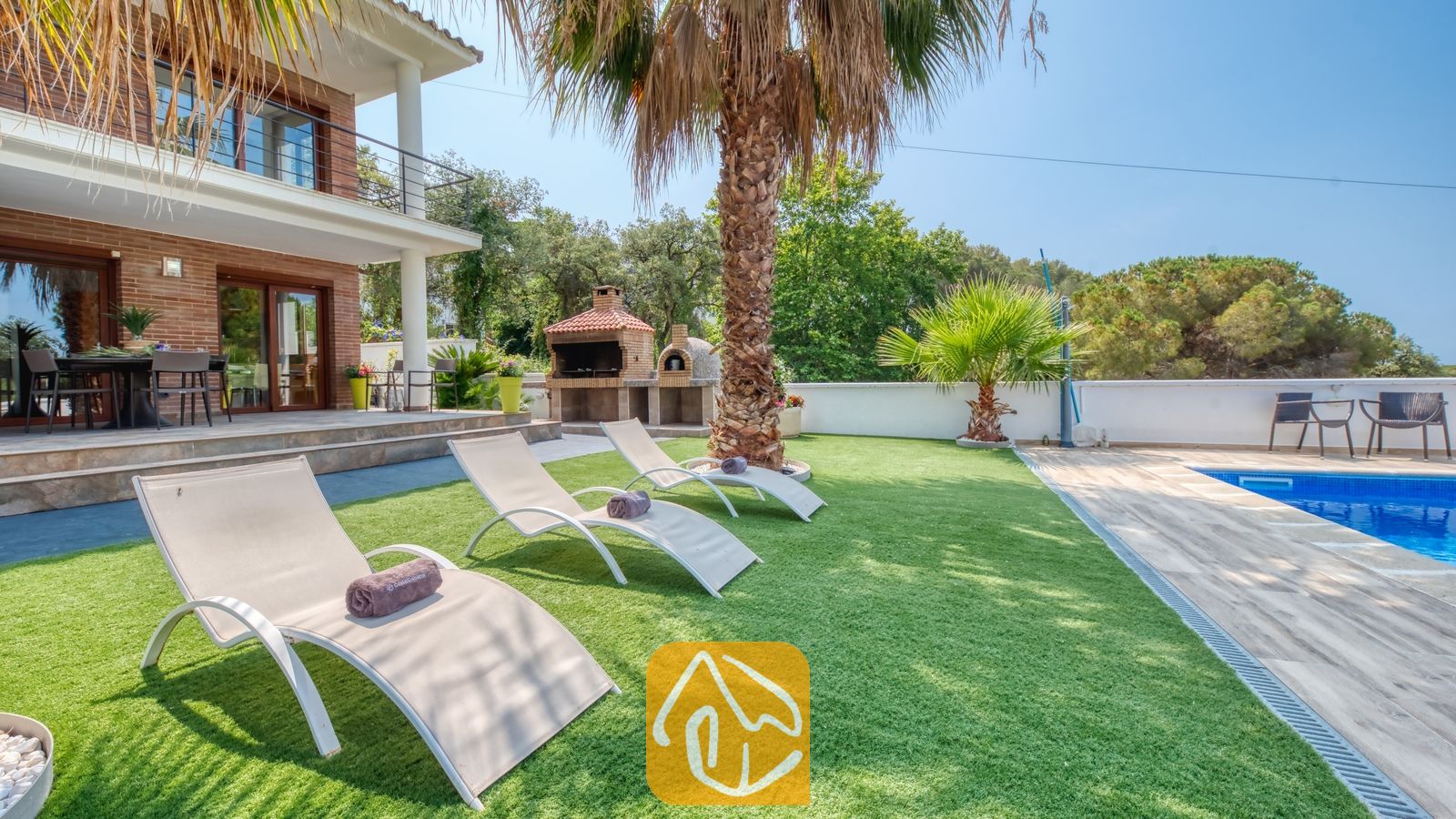 Vakantievilla's Costa Brava Spanje - Villa Marcella - Zonnige tuin bij Villa Marcella met witte ligstoelen op kunstgras, een palmboom en een blauw betegeld zwembad naast de bakstenen buitenkant van het huis.