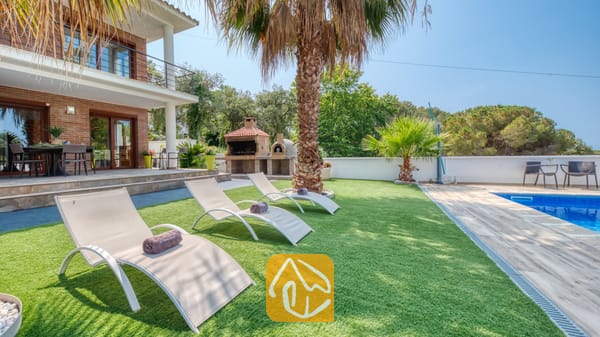 Vakantievilla's Costa Brava Spanje - Villa Marcella - Zonnige tuin bij Villa Marcella met witte ligstoelen op kunstgras, een palmboom en een blauw betegeld zwembad naast de bakstenen buitenkant van het huis.