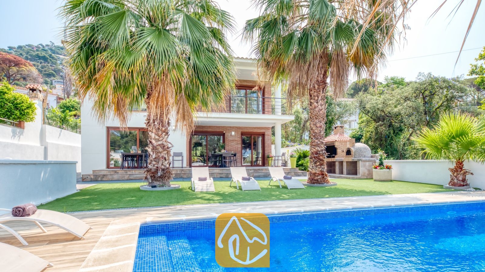 Vakantievilla's Costa Brava Spanje - Villa Marcella - Privézwembad met blauwe mozaïektegels en ligstoelen op het gras bij Villa Marcella, omgeven door palmbomen en voorzien van een stenen barbecue.