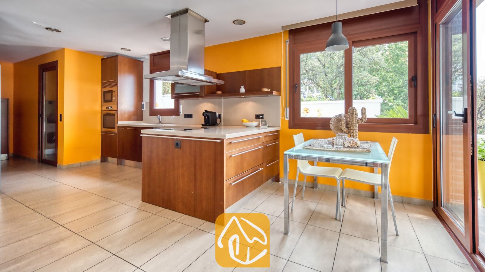 Vakantievilla's Costa Brava Spanje - Villa Marcella - Keuken met houten kasten, een kookeiland met stalen afzuigkap en een eettafel met witte stoelen tegen feloranje muren.