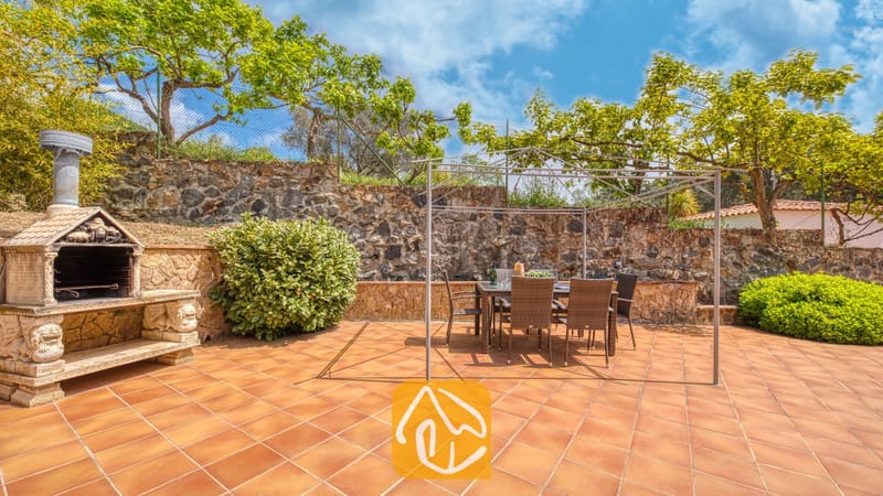 Vakantievilla's Costa Brava Spanje - Villa Lloret - Eethoek in de open lucht bij Villa Lloret, met een metalen pergola-frame boven een tafel met Wicker stoelen, gelegen op een betegeld terras naast een stenen barbecue.
