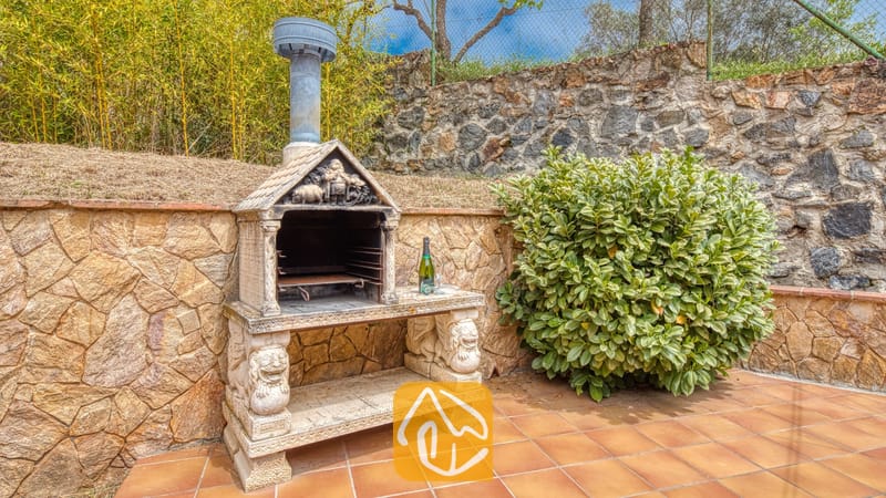 Vakantievilla's Costa Brava Spanje - Villa Lloret - Stenen buitenbarbecue met metalen schoorsteen op een betegeld terras naast een stenen keermuur en groene struik bij Villa Lloret.