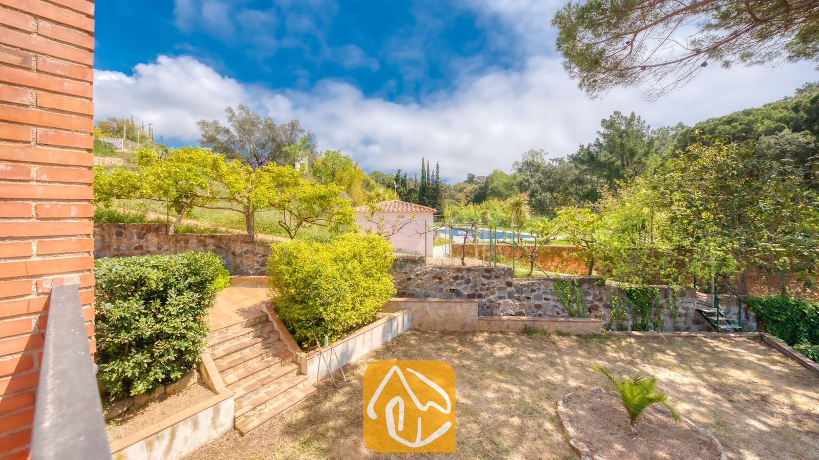 Vakantievilla's Costa Brava Spanje - Villa Lloret - Buitenaanzicht van Villa Lloret met aangelegde terrastuinen voorzien van stenen keermuren, betegelde buitentrappen en omringende groenbomen onder een blauwe lucht.