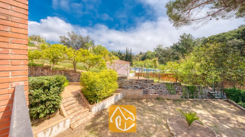 Vakantievilla's Costa Brava Spanje - Villa Lloret - Buitenaanzicht van Villa Lloret met aangelegde terrastuinen voorzien van stenen keermuren, betegelde buitentrappen en omringende groenbomen onder een blauwe lucht.