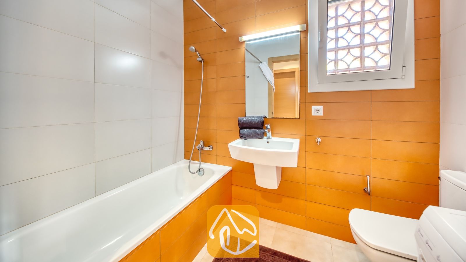 Vakantievilla's Costa Brava Spanje - Villa Lloret - Badkamer voorzien van een wit ligbad met handdouche, een wastafel met wandkraan onder een verlichte spiegel, en een raam met figuurglas tegen een achtergrond van oranje en witte wandtegels.