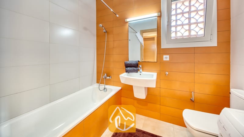 Vakantievilla's Costa Brava Spanje - Villa Lloret - Badkamer voorzien van een wit ligbad met handdouche, een wastafel met wandkraan onder een verlichte spiegel, en een raam met figuurglas tegen een achtergrond van oranje en witte wandtegels.