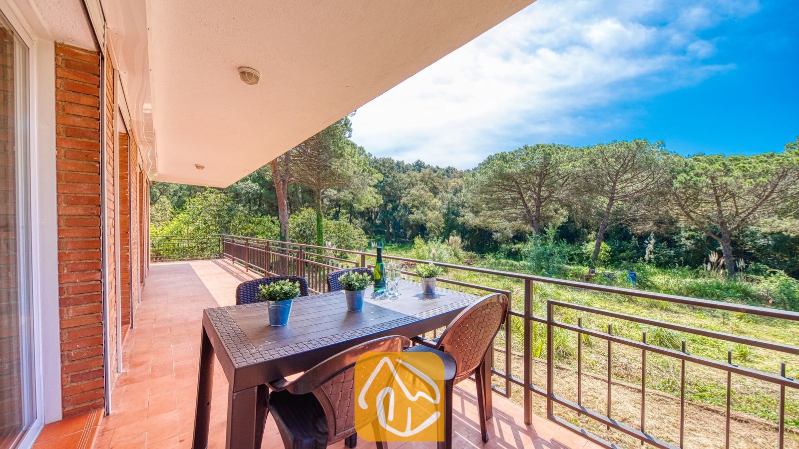 Vakantievilla's Costa Brava Spanje - Villa Lloret - Overdekt terras bij Villa Lloret met een bruine kunststof eettafel en stoelen, met uitzicht op groene dennenbomen en een blauwe lucht.