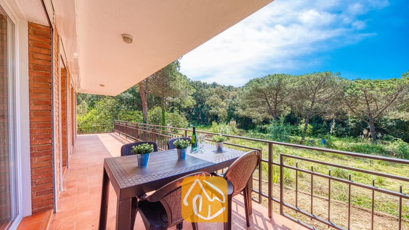 Vakantievilla's Costa Brava Spanje - Villa Lloret - Overdekt terras bij Villa Lloret met een bruine kunststof eettafel en stoelen, met uitzicht op groene dennenbomen en een blauwe lucht.