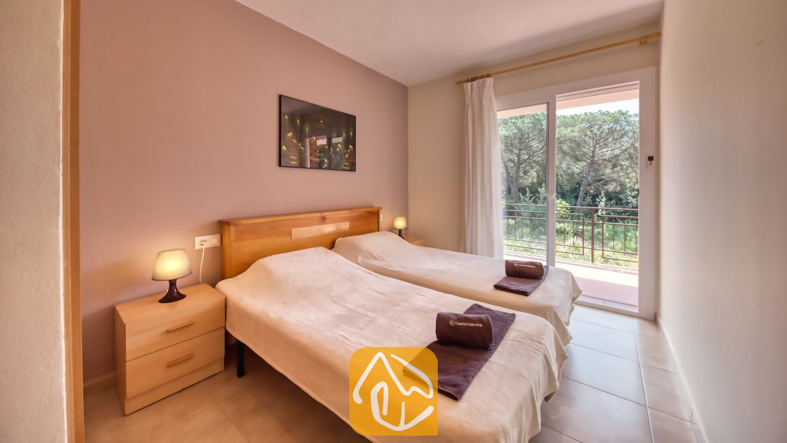 Vakantievilla's Costa Brava Spanje - Villa Lloret - Slaapkamer met twee eenpersoonsbedden met lichthouten frames, nachtkastjes met lampen, opgevouwen bruine handdoeken en een schuifpui die uitkomt op een balkon.