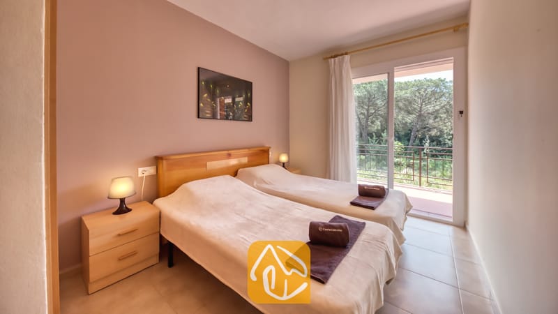 Vakantievilla's Costa Brava Spanje - Villa Lloret - Slaapkamer met twee eenpersoonsbedden met lichthouten frames, nachtkastjes met lampen, opgevouwen bruine handdoeken en een schuifpui die uitkomt op een balkon.
