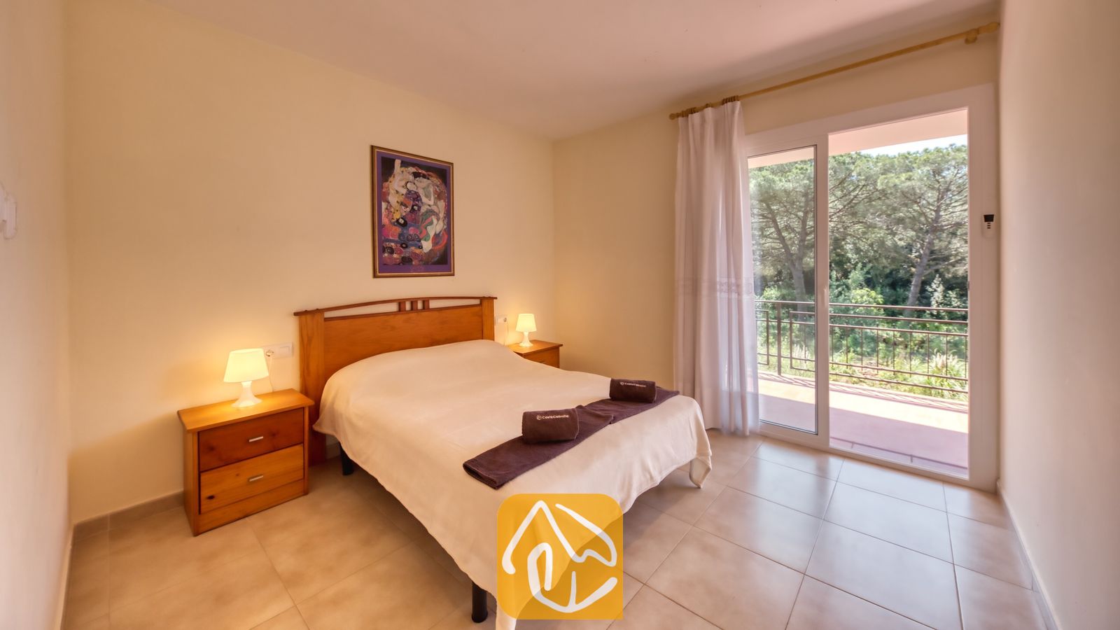 Vakantievilla's Costa Brava Spanje - Villa Lloret - Slaapkamer met een houten tweepersoonsbed, bijpassende nachtkastjes met lampen en glazen schuifdeuren die uitkomen op een balkon met bomen.