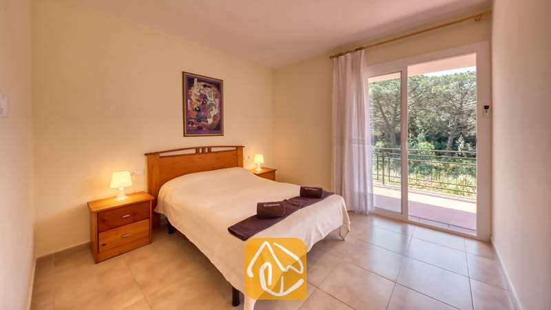 Vakantievilla's Costa Brava Spanje - Villa Lloret - Slaapkamer met een houten tweepersoonsbed, bijpassende nachtkastjes met lampen en glazen schuifdeuren die uitkomen op een balkon met bomen.