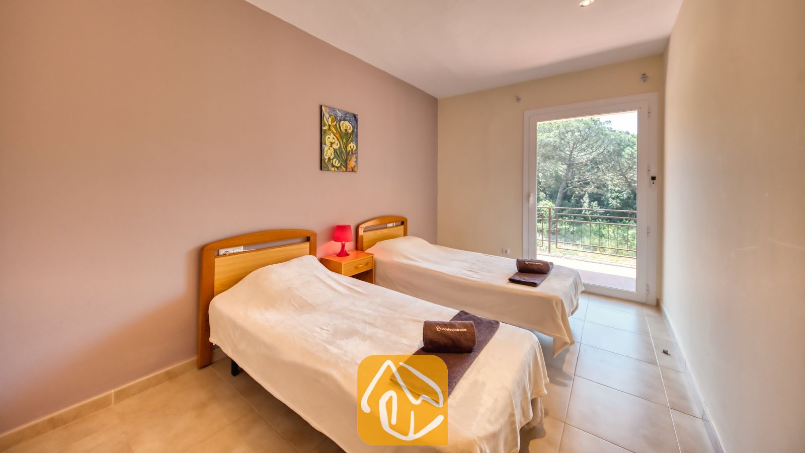Vakantievilla's Costa Brava Spanje - Villa Lloret - Slaapkamer met twee houten eenpersoonsbedden, witte spreuken, opgerolde bruine handdoeken, een klein nachtkastje met een rode lamp en een glazen deur die uitkomt op een balkon met bomen.