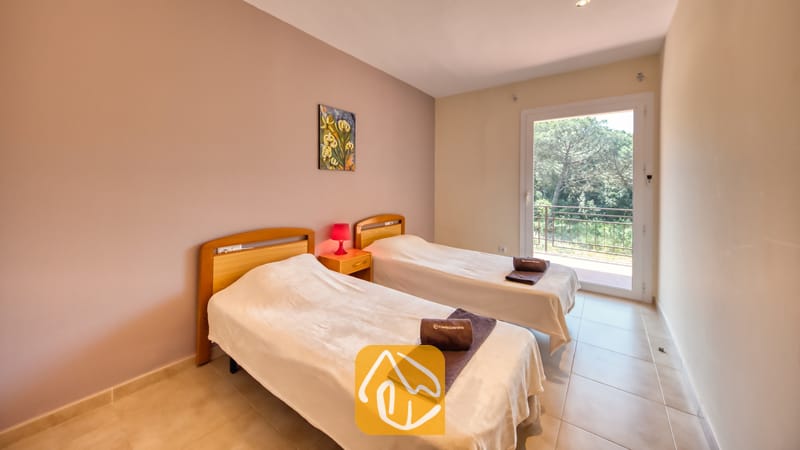 Vakantievilla's Costa Brava Spanje - Villa Lloret - Slaapkamer met twee houten eenpersoonsbedden, witte spreuken, opgerolde bruine handdoeken, een klein nachtkastje met een rode lamp en een glazen deur die uitkomt op een balkon met bomen.