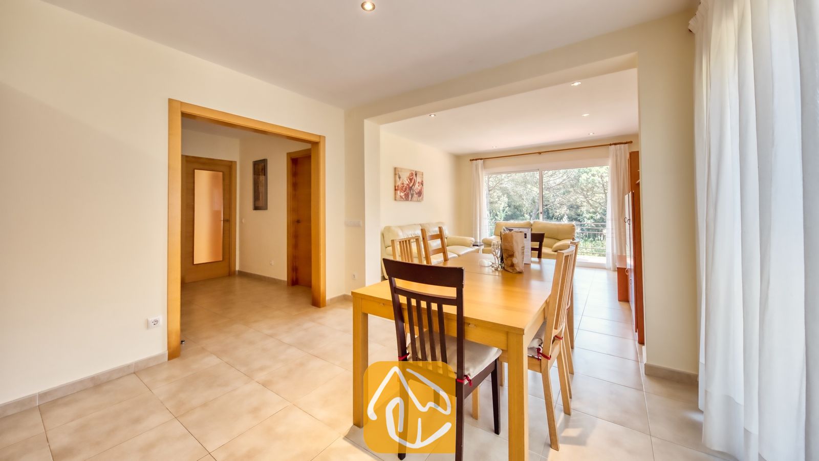 Vakantievilla's Costa Brava Spanje - Villa Lloret - Houten eettafel met stoelen op de voorgrond van het woongedeelte in Villa Lloret, met tegelvloeren en een deuropening die leidt naar de gang.
