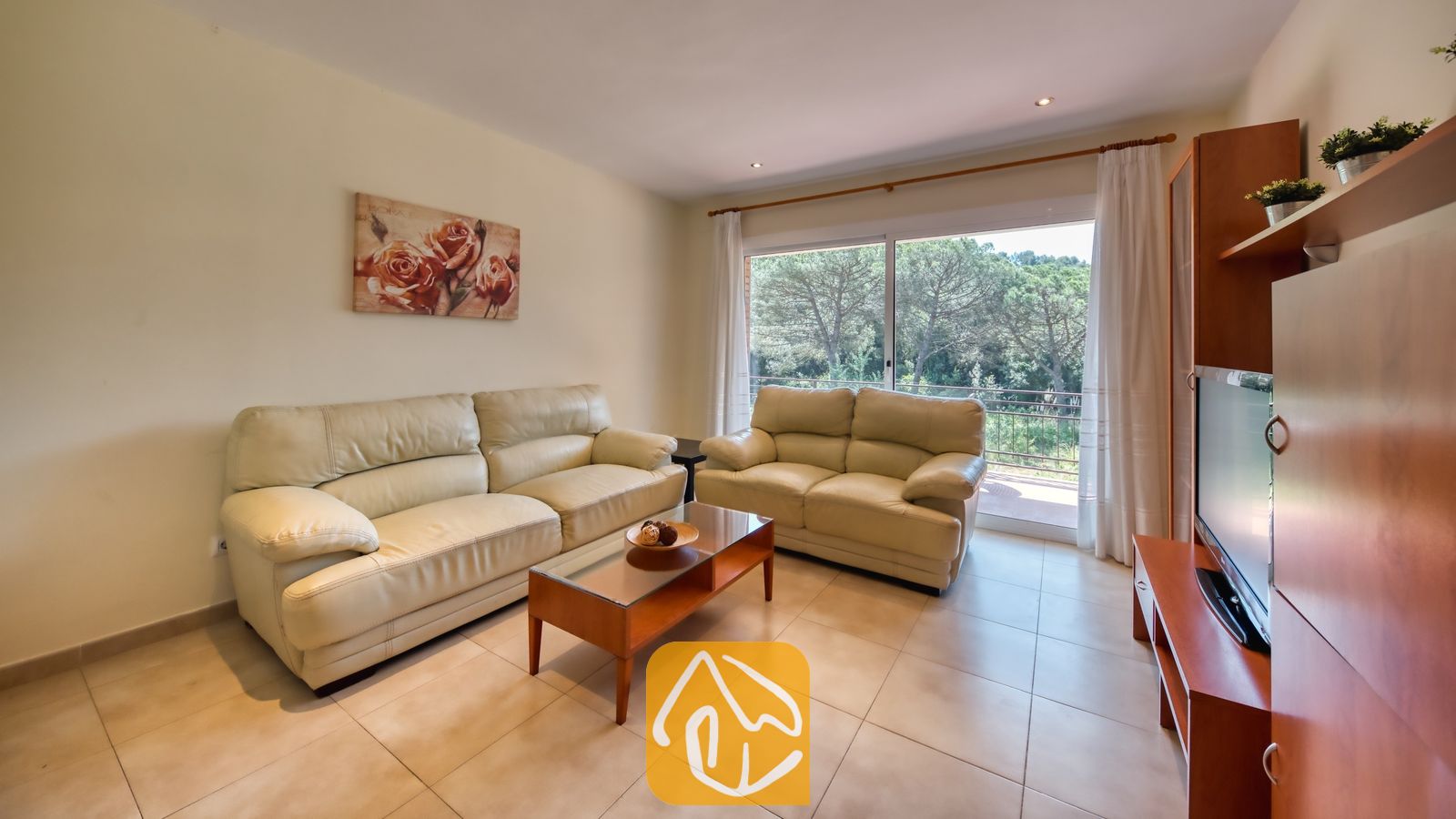 Vakantievilla's Costa Brava Spanje - Villa Lloret - Woonkamer met twee crèmelederen banken, een houten salontafel met glazen blad en een tv-meubel in Villa Lloret, voorzien van een groot raam met uitzicht op groene bomen.