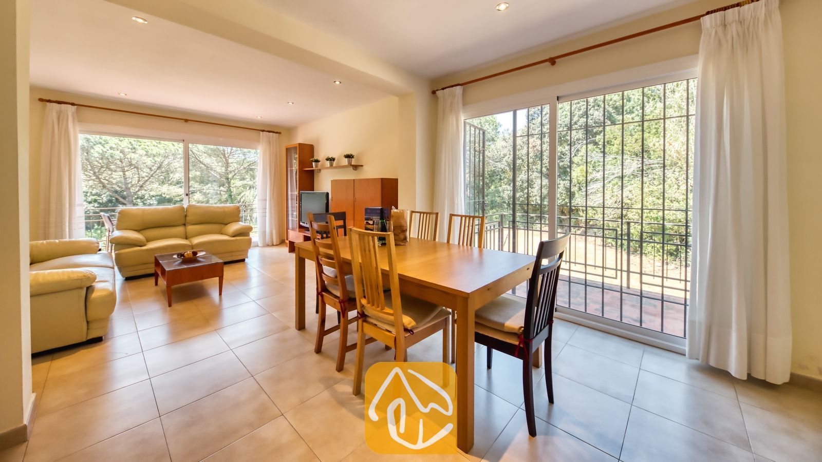 Vakantievilla's Costa Brava Spanje - Villa Lloret - Ruime, open woon- en eetkamer in Villa Lloret met een houten eettafel, crèmelederen banken, tegelvloeren en grote glazen schuifpuien die uitkomen op een balkon met groene bomen.