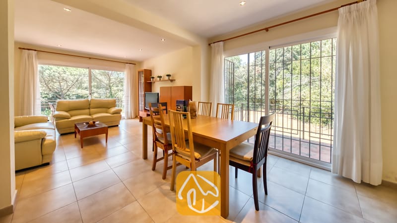 Vakantievilla's Costa Brava Spanje - Villa Lloret - Ruime, open woon- en eetkamer in Villa Lloret met een houten eettafel, crèmelederen banken, tegelvloeren en grote glazen schuifpuien die uitkomen op een balkon met groene bomen.
